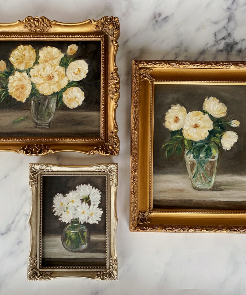 May include: Three framed paintings of white flowers in vases. The paintings are in gold and silver frames. The flowers are in different stages of bloom.
