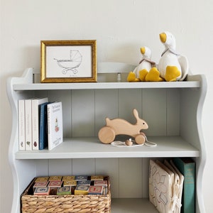 May include: A white wooden bookshelf with three shelves. The top shelf has a gold framed print of a baby carriage, and two white stuffed geese with yellow beaks. The middle shelf has a wooden toy bunny with wheels, and a stack of books. The bottom shelf has a wicker basket filled with colorful wooden blocks, a white fabric book with geometric shapes, and a green book titled "Anne of Green Gables".