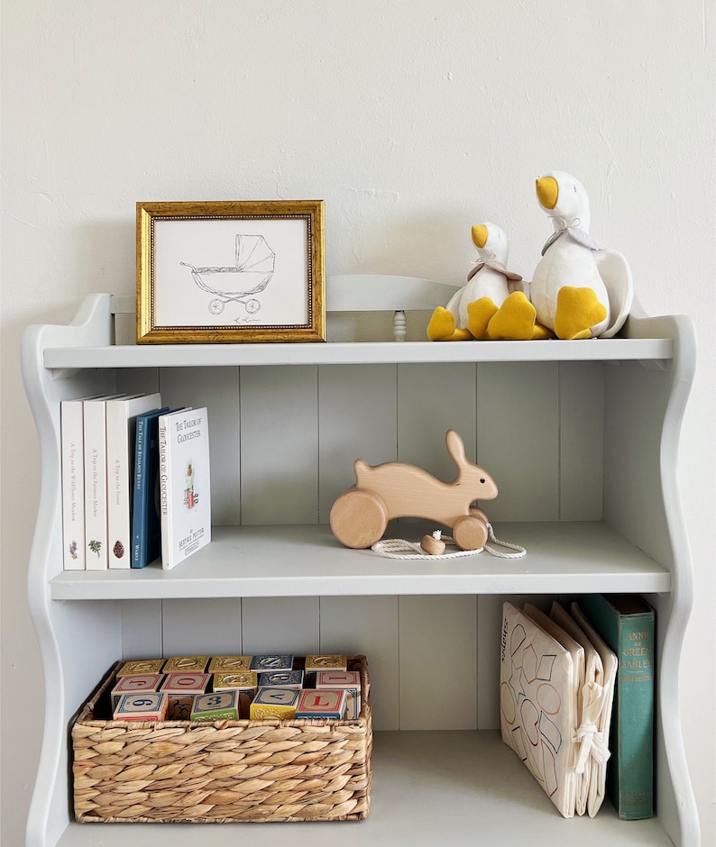May include: A white wooden bookshelf with three shelves. The top shelf has a gold framed print of a baby carriage, and two white stuffed geese with yellow beaks. The middle shelf has a wooden toy bunny with wheels, and a stack of books. The bottom shelf has a wicker basket filled with colorful wooden blocks, a white fabric book with geometric shapes, and a green book titled "Anne of Green Gables".