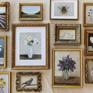 May include: A gallery wall of framed artwork featuring floral, animal, and nature scenes. The frames are all gold and have ornate designs. The artwork includes a bee, a chickadee, a bouquet of white flowers, a bouquet of purple flowers, a bouquet of white daisies, and a field of lavender.