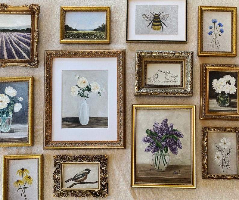 May include: A gallery wall of framed artwork featuring floral, animal, and nature scenes. The frames are all gold and have ornate designs. The artwork includes a bee, a chickadee, a bouquet of white flowers, a bouquet of purple flowers, a bouquet of white daisies, and a field of lavender.