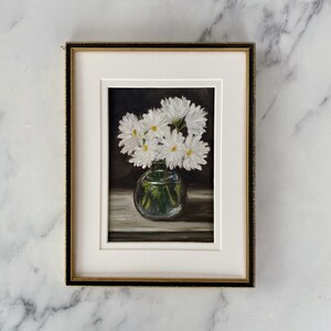 May include: A framed painting of a bouquet of white daisies in a clear glass vase on a dark background.