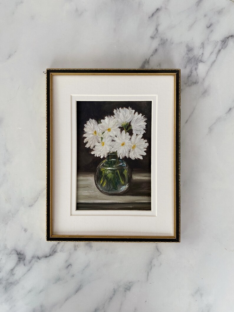 May include: A framed painting of a bouquet of white daisies in a clear glass vase on a dark background.