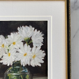 May include: A framed painting of a bouquet of white daisies in a clear glass vase. The flowers are arranged in a cluster and the vase is sitting on a table. The background is dark.