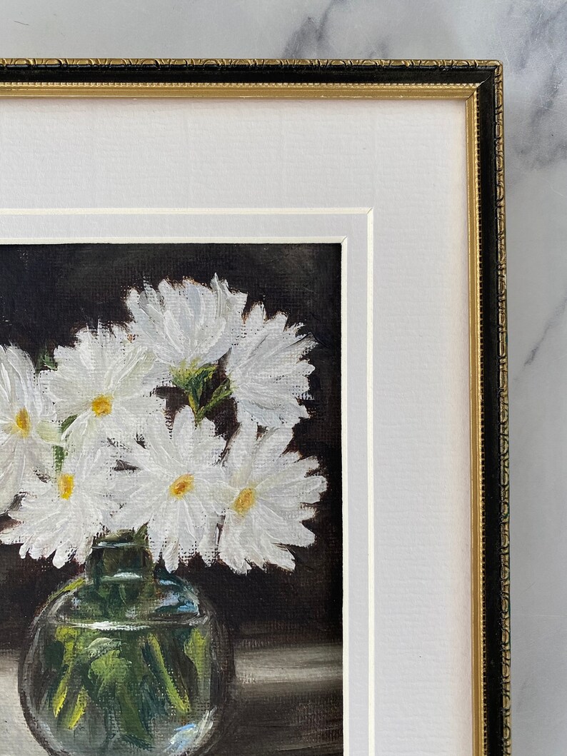 May include: A framed painting of a bouquet of white daisies in a clear glass vase. The flowers are arranged in a cluster and the vase is sitting on a table. The background is dark.