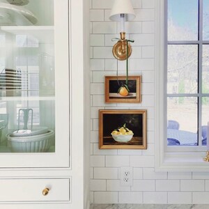 May include: A white kitchen with white subway tile backsplash, a gold light fixture, and two framed still life paintings of fruit. The paintings are hung above a white electrical outlet.