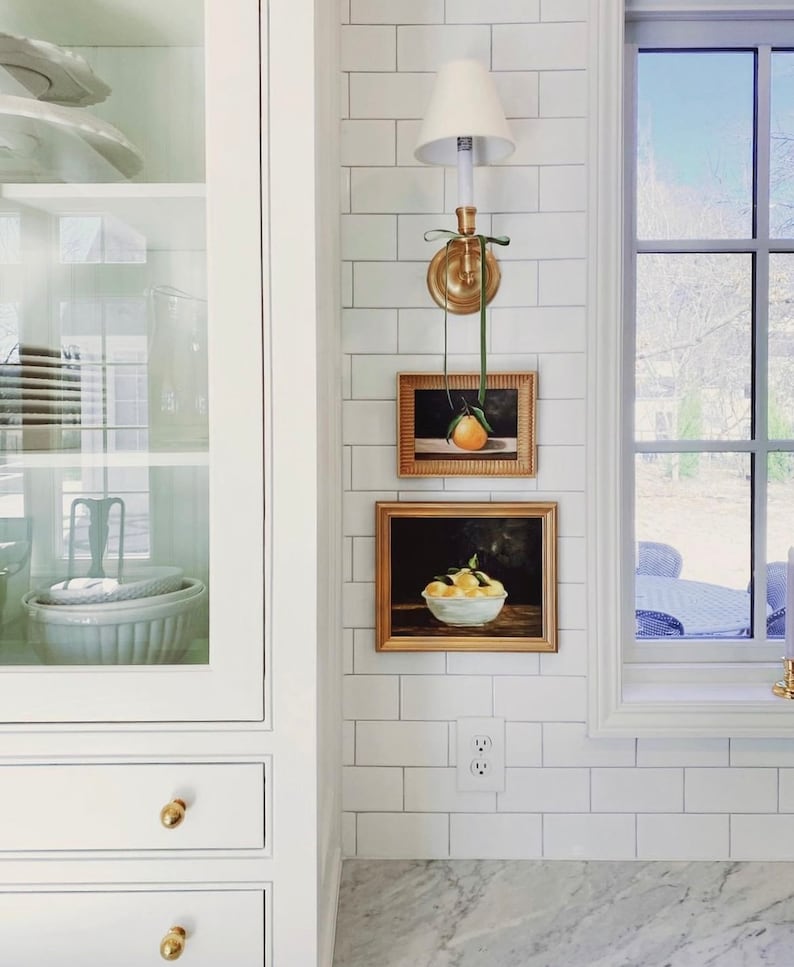 May include: A white kitchen with white subway tile backsplash, a gold light fixture, and two framed still life paintings of fruit. The paintings are hung above a white electrical outlet.