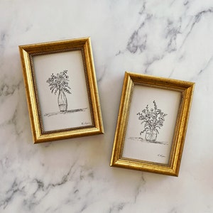May include: Two gold-framed prints of black and white line drawings of flower arrangements in vases. The prints are on white paper and have the artist's signature in the bottom right corner.