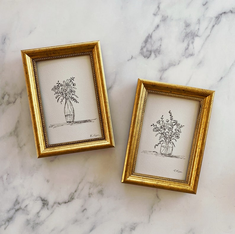 May include: Two gold-framed prints of black and white line drawings of flower arrangements in vases. The prints are on white paper and have the artist's signature in the bottom right corner.