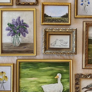 May include: A wall gallery of framed artwork featuring various subjects, including a duck, flowers, and landscapes. The frames are all gold or silver and have ornate details.