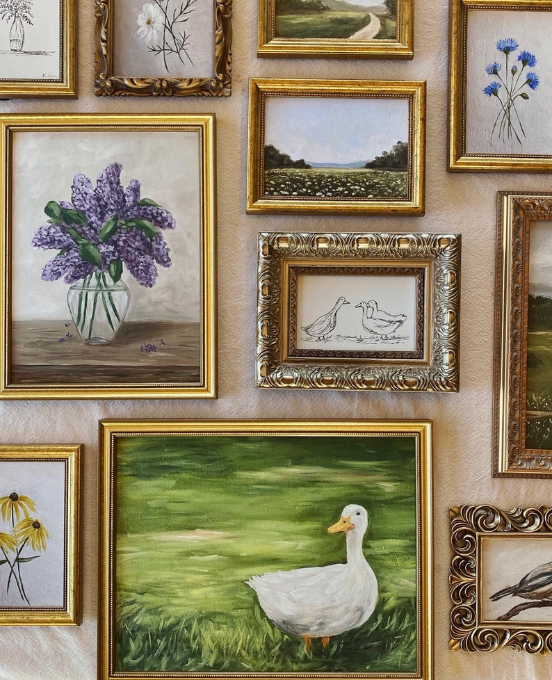 May include: A wall gallery of framed artwork featuring various subjects, including a duck, flowers, and landscapes. The frames are all gold or silver and have ornate details.