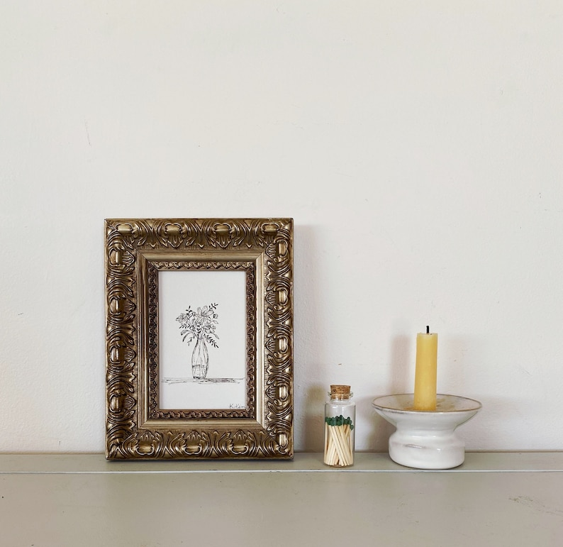 May include: A black and white line drawing of flowers in a vase is framed in a gold ornate frame. A small glass jar filled with matches and a white ceramic candle holder with a yellow candle are on a shelf.