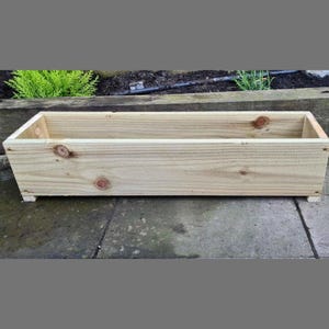 May include: A rectangular wooden planter box made from light-coloured wood. The box has a simple design with visible wood grain and knots. It sits on small feet, ready for planting flowers or herbs.