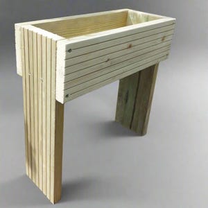 May include: A wooden planter box, constructed from light-coloured timber. The rectangular box has horizontal slats and is supported by two vertical legs and a slanted support. The planter is designed for outdoor use, perfect for gardens and patios.