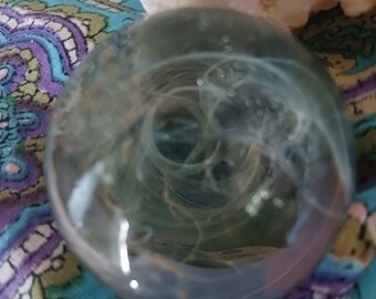 May include: A clear glass sphere with a swirling, cloudy pattern inside. The sphere is resting on a colorful patterned fabric.