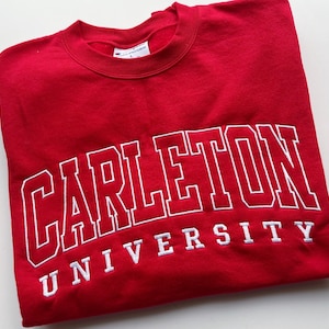 May include: A red sweatshirt featuring the white embroidered text "CARLETON UNIVERSITY". The sweatshirt is a Champion brand, size L. The garment is folded neatly.