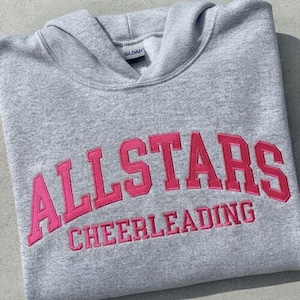Puede incluir: Una sudadera con capucha de color gris claro que presenta el texto ALLSTARS CHEERLEADING bordado en letras de bloque de color rosa brillante en el pecho. La prenda incluye una capucha y una etiqueta de la marca Gildan en el cuello.