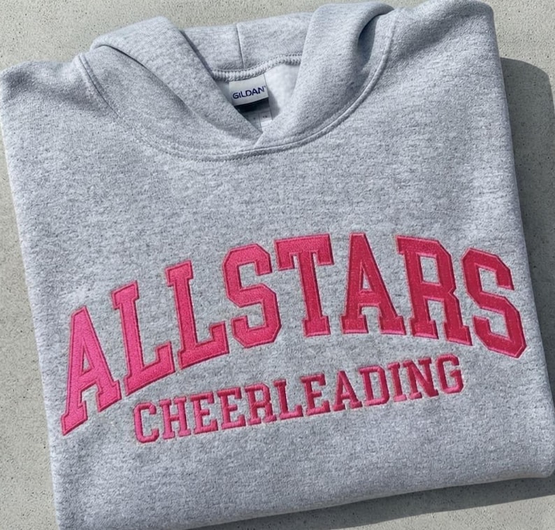 Puede incluir: Una sudadera con capucha de color gris claro que presenta el texto ALLSTARS CHEERLEADING bordado en letras de bloque de color rosa brillante en el pecho. La prenda incluye una capucha y una etiqueta de la marca Gildan en el cuello.