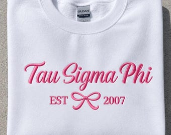 May include: A white crewneck sweatshirt featuring pink embroidered text that reads Tau Sigma Phi. Below the text, the word EST is followed by a pink embroidered bow icon and the number 2007. The garment has a Gildan brand label at the neck.
