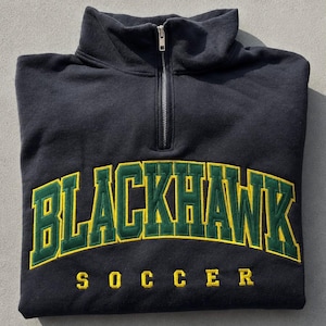 K&ouml;nnte beinhalten: Ein schwarzes Sweatshirt mit kurzem Rei&szlig;verschluss am Kragen, das den Schriftzug BLACKHAWK in gro&szlig;en gr&uuml;nen Buchstaben mit gelben Umrandungen bogenf&ouml;rmig auf der Brust zeigt. Darunter steht SOCCER in gelben Buchstaben.