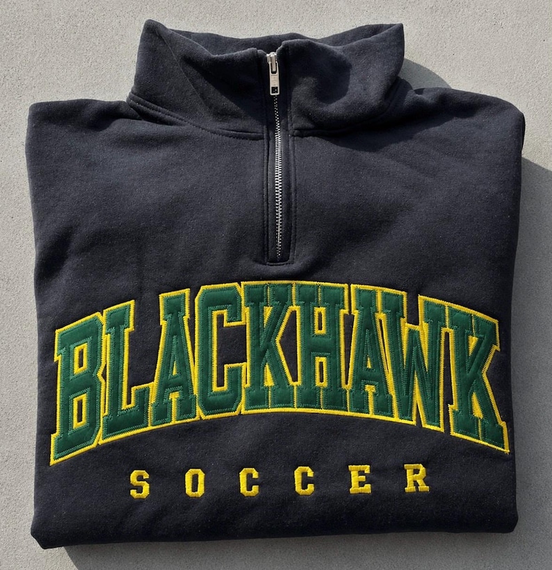 K&ouml;nnte beinhalten: Ein schwarzes Sweatshirt mit kurzem Rei&szlig;verschluss am Kragen, das den Schriftzug BLACKHAWK in gro&szlig;en gr&uuml;nen Buchstaben mit gelben Umrandungen bogenf&ouml;rmig auf der Brust zeigt. Darunter steht SOCCER in gelben Buchstaben.
