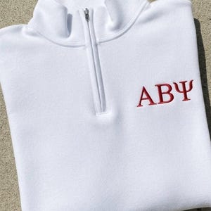 Peut inclure: Un pull blanc à fermeture éclair quart de tour avec un col montant et une fermeture éclair en métal argenté. Le vêtement présente les lettres grecques Alpha, Bêta et Psi brodées en fil rouge sur la poitrine supérieure droite.