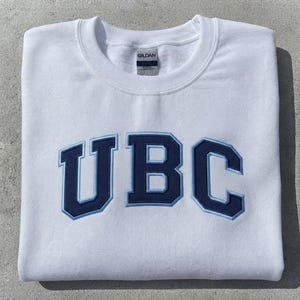 May include: A white crewneck sweatshirt featuring the letters UBC embroidered in dark navy blue with a light blue outline across the chest. The garment has a ribbed collar and a Gildan brand label visible at the inside of the neckline.