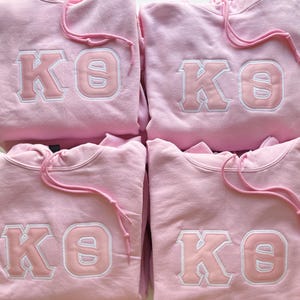 May include: Four light pink hooded sweatshirts featuring the Greek letters Kappa and Theta embroidered in a lighter shade of pink with a white border. Each hoodie includes a matching pink drawstring at the neckline and a front kangaroo pocket.