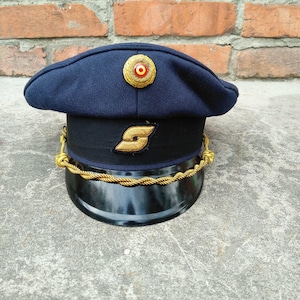 May include: A navy blue peaked cap featuring a black band with a gold-colored emblem, a gold braided chin strap, and a circular gold-toned badge with a red and yellow center. The cap has a black visor and a structured crown made of dark blue fabric.