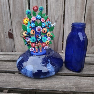 May include: A blue ceramic bottle stopper with a colorful ceramic tree of life design. The tree features flowers, butterflies, a sun, a moon, and two figures. The bottle stopper is sitting on a wooden surface.