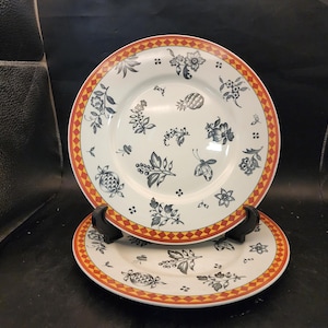 May include: Two white ceramic plates with a floral design. The plates have a red and orange diamond pattern border. The plates are decorated with black and gray floral and fruit motifs.