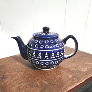 May include: A round ceramic teapot featuring a deep blue glaze with white hand-painted patterns. The design includes horizontal bands of small star-like flowers and a central row of stylized evergreen trees. It has a matching lid and handle.