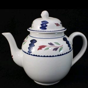 May include: White ceramic teapot with a lid, spout, and handle. The teapot is decorated with blue, red, and green floral patterns and a scalloped blue border. The lid has a small knob.