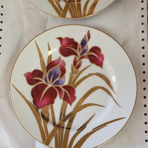 May include: Set of decorative plates featuring a floral design. Each plate has a white background with a burgundy and purple iris flower and gold accents. The plates have a gold rim.