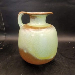 May include: A ceramic pitcher with a rounded body and a single handle. The surface features a two-tone glaze with a pale mint green color on the upper portion and a warm brown tone on the lower section, blending together in a soft gradient.