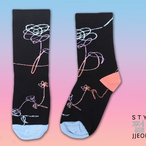 May include: A pair of black socks with a light pink and blue floral design. The socks have a light blue cuff and a light pink heel and toe. The socks feature the text "STYLE 쩔어 JJEOREO".