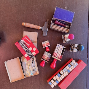 Op de afbeelding: Een verzameling vintage kantoorbenodigdheden, waaronder stempels, een hamer en dozen met bevestigingsmiddelen. De afbeelding toont een rood-witte doos met "PRESTONG" papieren bevestigingsmiddelen, samen met diverse stempels met rode handvatten en een blauwe doos.