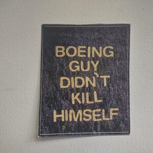 Pode incluir: Um adesivo preto e branco com o texto "BOEING GUY DIDN'T KILL HIMSELF" em letras douradas.