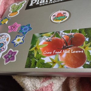 May include: A sticker with a close-up of red tomatoes on a vine with green leaves and the text "Grow Food Not Lawns".
