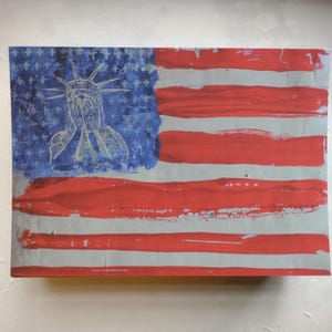 May include: A rectangular artwork depicting the American flag with a distressed, painted aesthetic. The flag features red and white stripes, a blue field with white stars, and a sketched image of the Statue of Liberty.