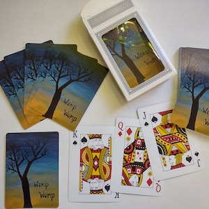 Puede incluir: Una baraja de cartas con diseños personalizados en el dorso que representan una puesta de sol azul y amarilla con la silueta de un árbol. El texto "Womp Womp" está impreso en las cartas. Las cartas están dispuestas sobre una superficie blanca con una caja de cartas en el fondo.