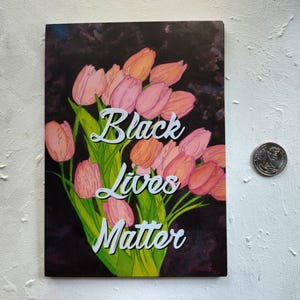 May include: An art print showing a bouquet of pink and orange tulips on a dark backdrop. The words "Black Lives Matter" are in white script. A coin is next to the print. The print is rectangular.