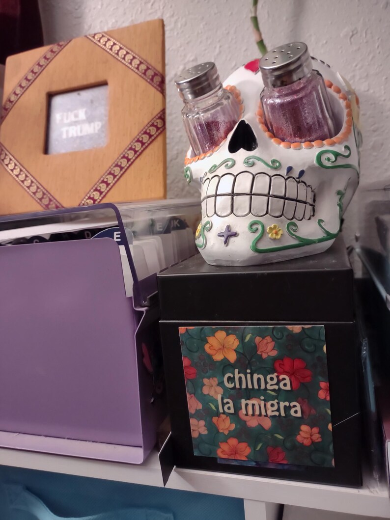 Puede incluir: Un juego de salero y pimentero de calavera de az&uacute;car blanca con un dise&ntilde;o floral. La calavera tiene un dise&ntilde;o floral verde y amarillo y un coraz&oacute;n rojo en la frente. Los saleros y pimenteros est&aacute;n en las cuencas de los ojos. La calavera est&aacute; sentada sobre una caja negra con una pegatina que dice "chinga la migra" en un dise&ntilde;o floral.