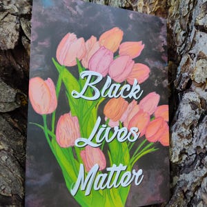 May include: A card displaying a floral arrangement of pink and orange tulips with green foliage, set against a dark backdrop. The words "Black Lives Matter" are written in white script. The card is shown against a textured tree trunk.