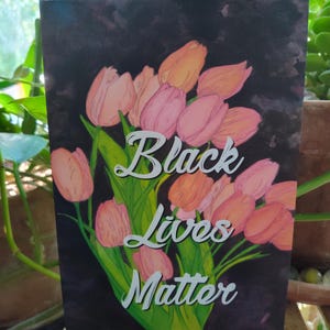 May include: A print with a dark background, displaying a bouquet of pink and peach tulips. The words "Black Lives Matter" are written in white script. Green leaves and stems are also visible in the image.