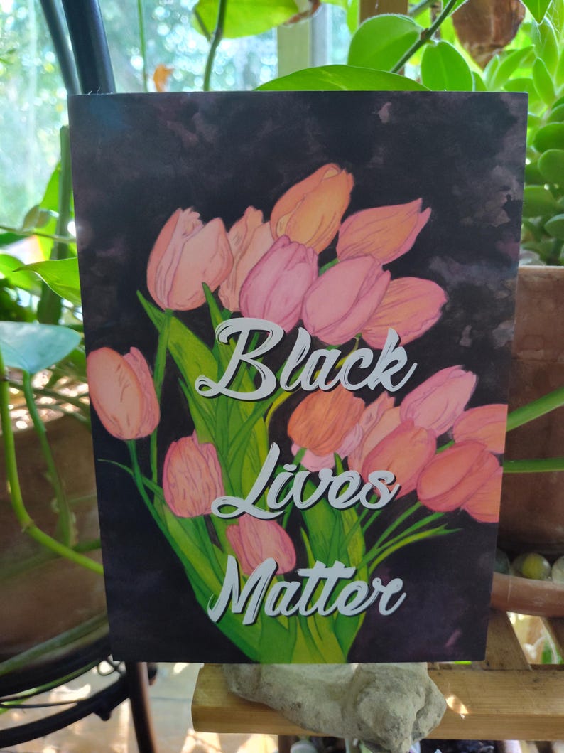 May include: A print with a dark background, displaying a bouquet of pink and peach tulips. The words "Black Lives Matter" are written in white script. Green leaves and stems are also visible in the image.