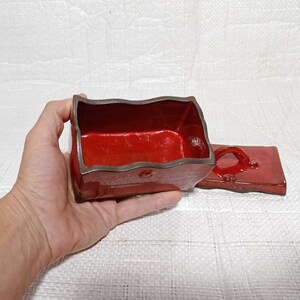 May include: A red ceramic box with a lid. The box has a wavy edge and a brown rim. The lid has a handle.