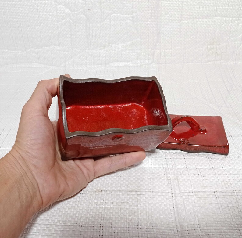May include: A red ceramic box with a lid. The box has a wavy edge and a brown rim. The lid has a handle.