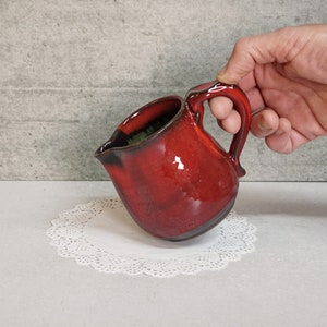 Könnte beinhalten: Ein roter Keramik-Krug mit einem dunkelbraunen Rand und einem Henkel. Der Krug steht auf einer weißen Spitze.