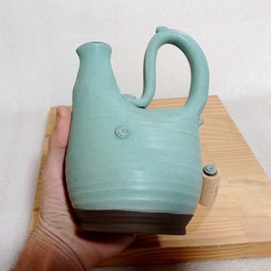 May include: A teal blue ceramic pitcher with a unique, curved handle. The pitcher has a textured surface and a dark brown base.